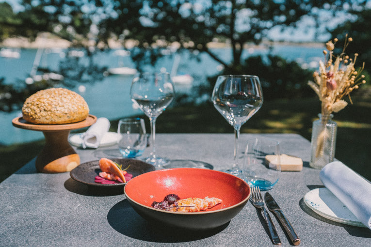 Table dressée en extérieur avec vue sur un plan d'eau, comprenant un plat élégant dans une assiette rouge, du pain sur un support en bois et des verres à vin prêts pour un repas raffiné.