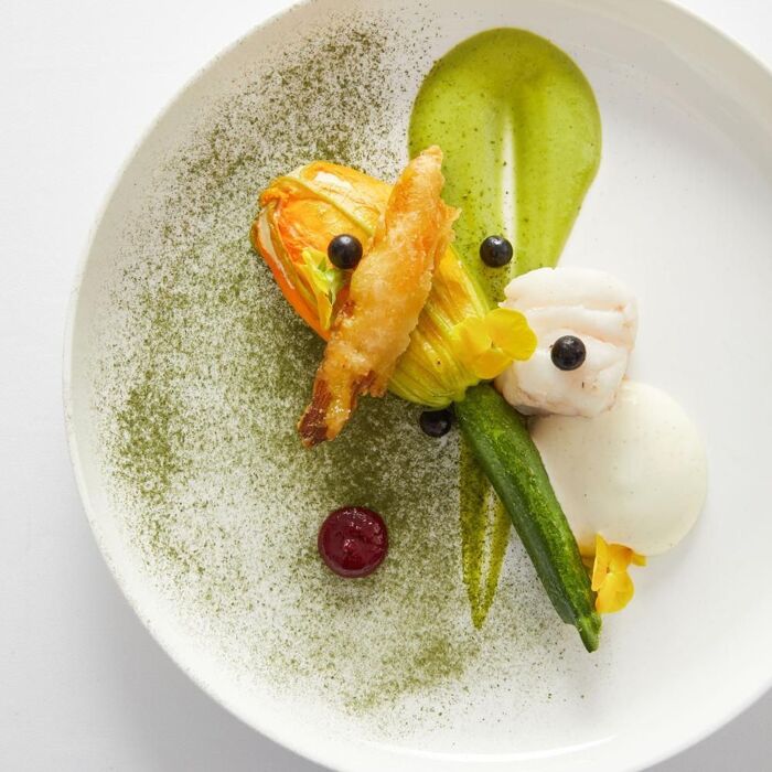Fleur de courgette farcie et autres ingrédients délicats, artistiquement disposés sur une assiette blanche avec une sauce verte.

