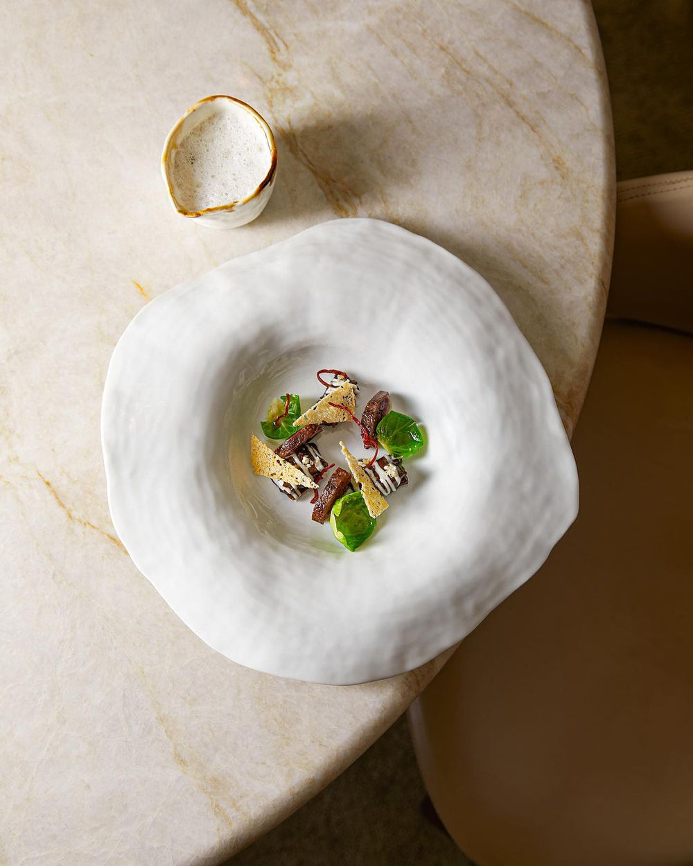 Plat gastronomique dans une assiette blanche texturée avec des légumes et des garnitures artistiques, accompagné d'un petit bol de sauce à côté.