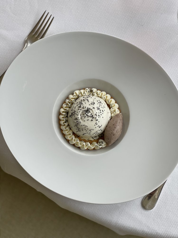 Dessert dans une assiette blanche, composé d'une crème fouettée garnie de graines de pavot, entourée d'une décoration de crème et accompagnée d'une quenelle de glace.