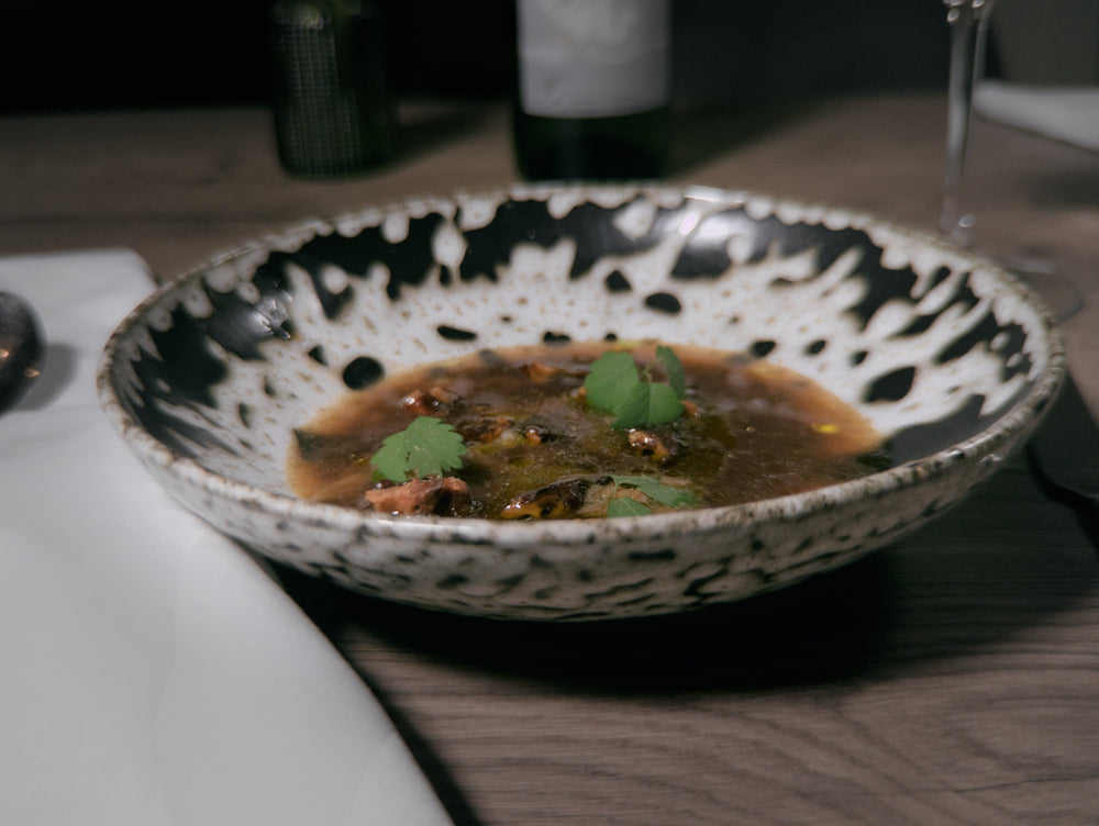 Plat gastronomique présenté dans une assiette en céramique blanche et noire, contenant une soupe ou un ragoût avec des morceaux de viande et des herbes fraîches. La table est dressée avec une serviette blanche et un verre à vin en arrière-plan. L'ambiance est élégante et raffinée.