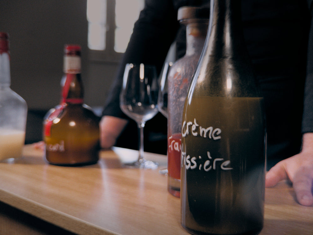 Gros plan sur des bouteilles de liqueur disposées sur un comptoir en bois, avec un verre à vin vide au centre. Une des bouteilles est étiquetée 
