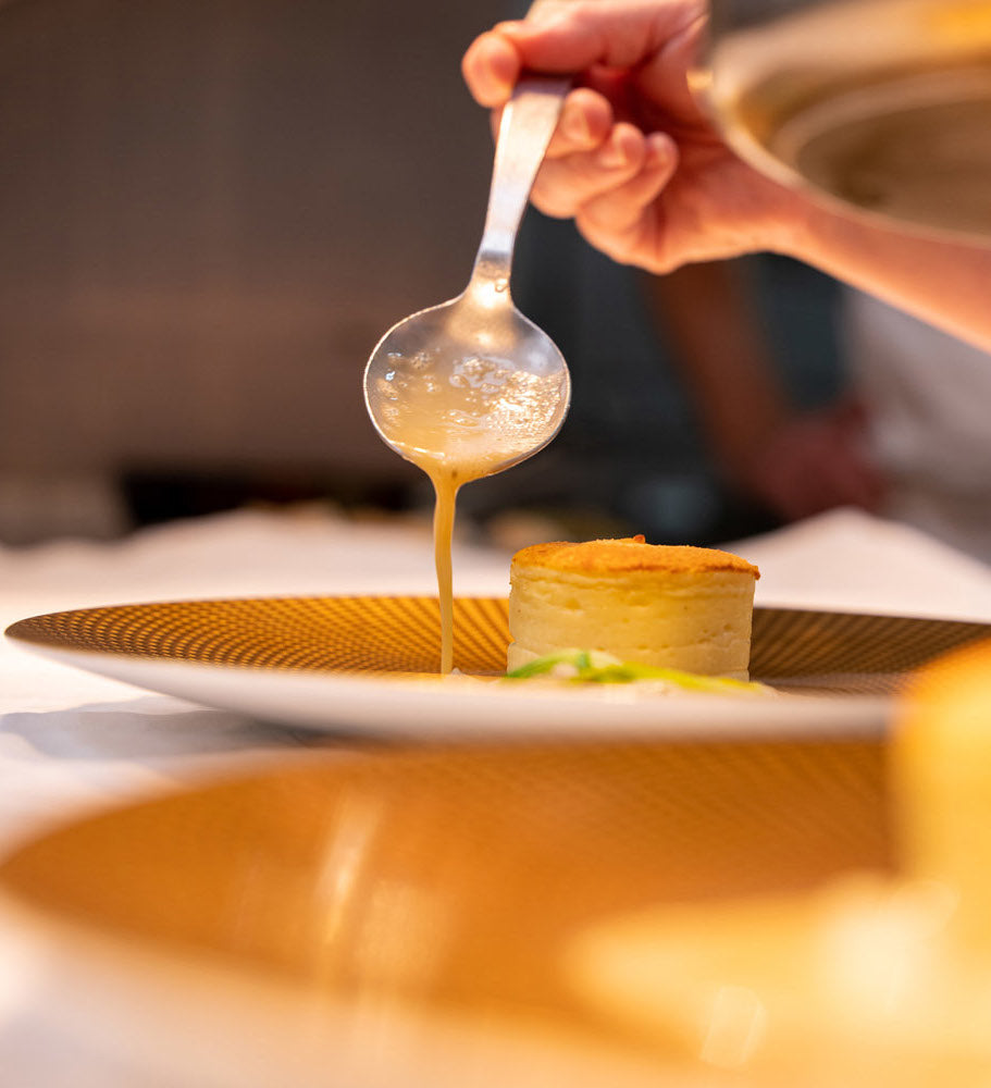 Une cuillère versant une sauce délicate sur un soufflé de pommes de terre servi dans une assiette dorée.