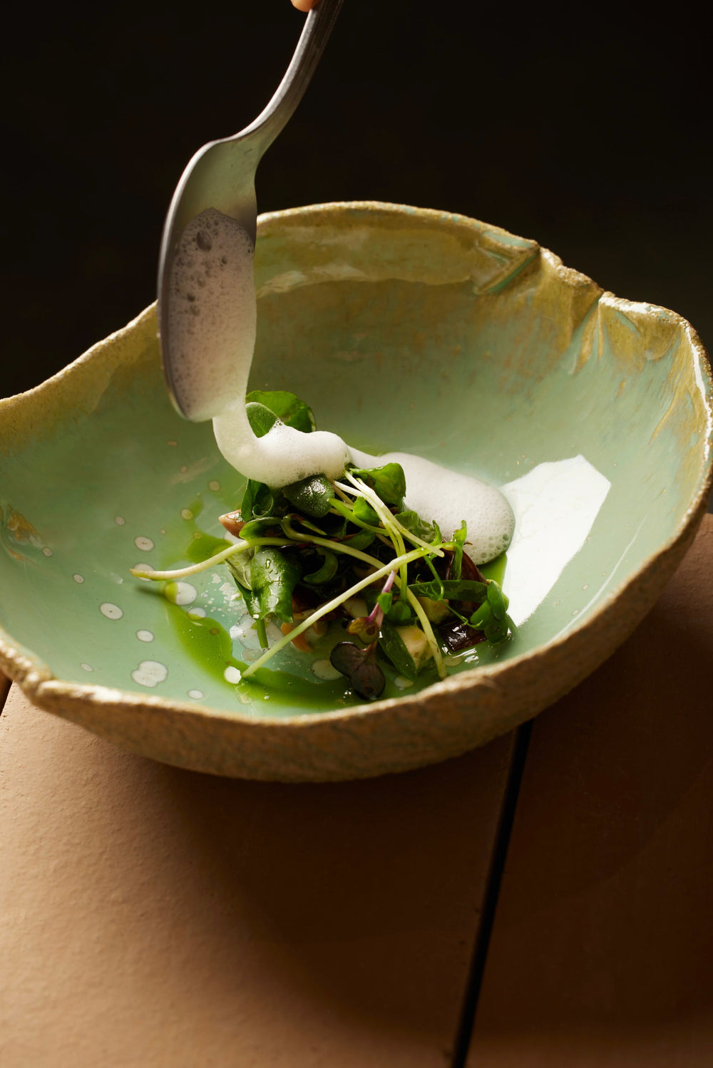 Un plat gastronomique délicatement préparé, garni de jeunes pousses et arrosé d'une émulsion crémeuse versée avec une cuillère dans un bol en céramique verte.