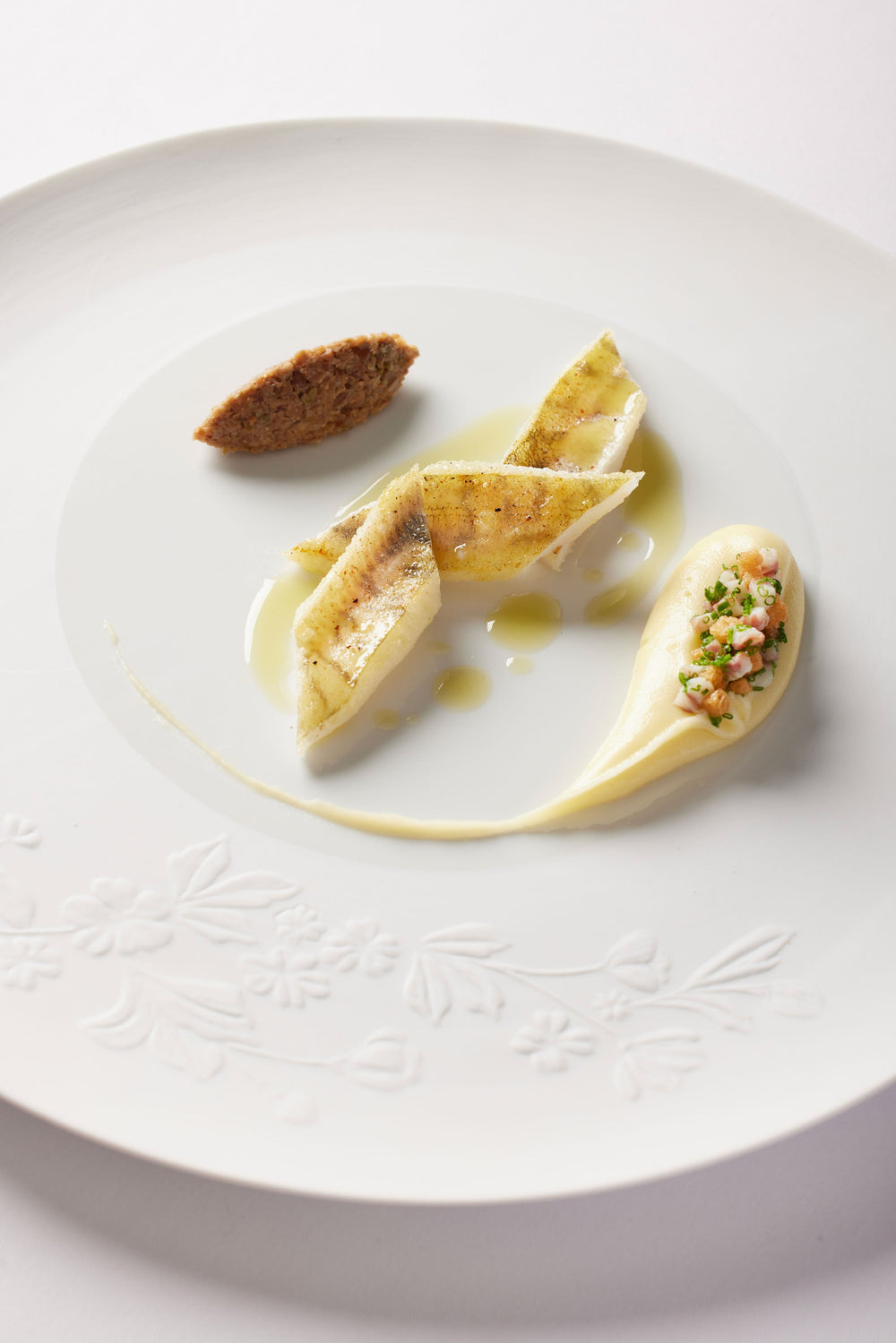 Assiette gastronomique élégante avec un plat de poisson délicatement dressé, accompagné d'une quenelle de purée et d'une garniture raffinée, sur une assiette blanche ornée de motifs floraux.