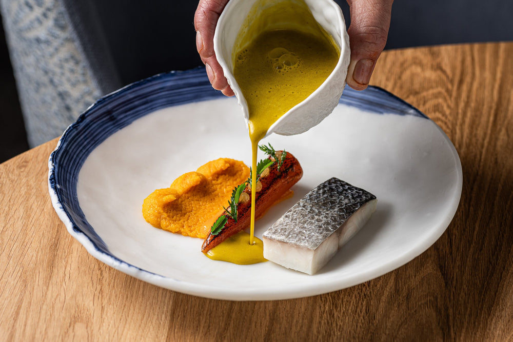 Plat gastronomique avec sauce jaune versée sur un poisson et des légumes, plat présenté dans une assiette bleue.