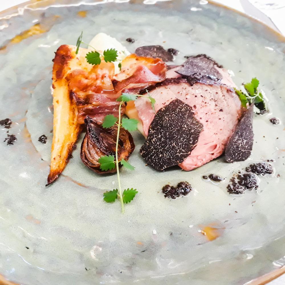 Un plat gastronomique où une tranche de viande est agrémentée de truffes, d’oignons rôtis, et d’autres légumes, joliment disposés sur une assiette en céramique.