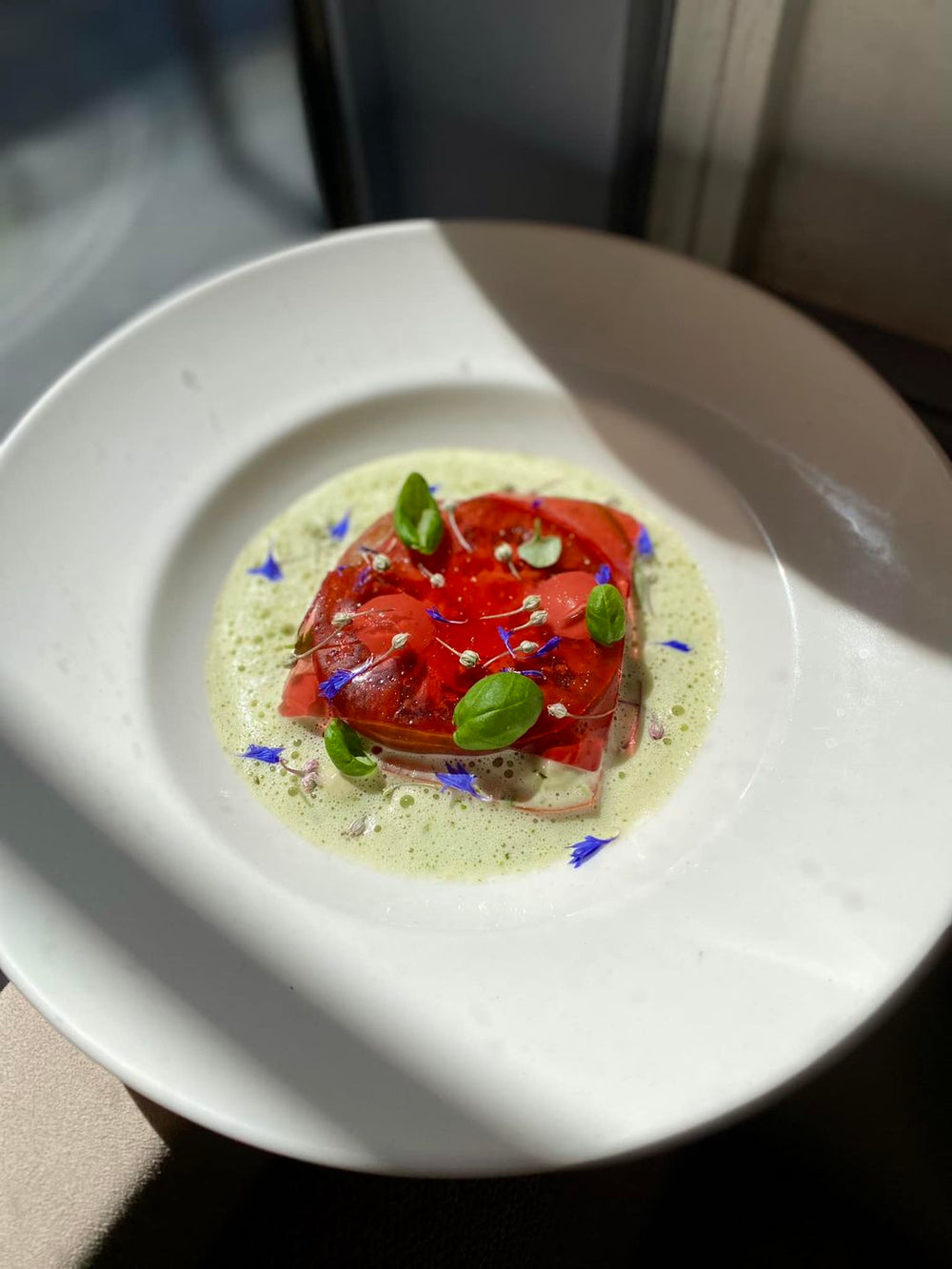 Carpaccio de tomates avec mousse verte et décor floral, servi dans une assiette blanche épurée baignée de lumière naturelle.
