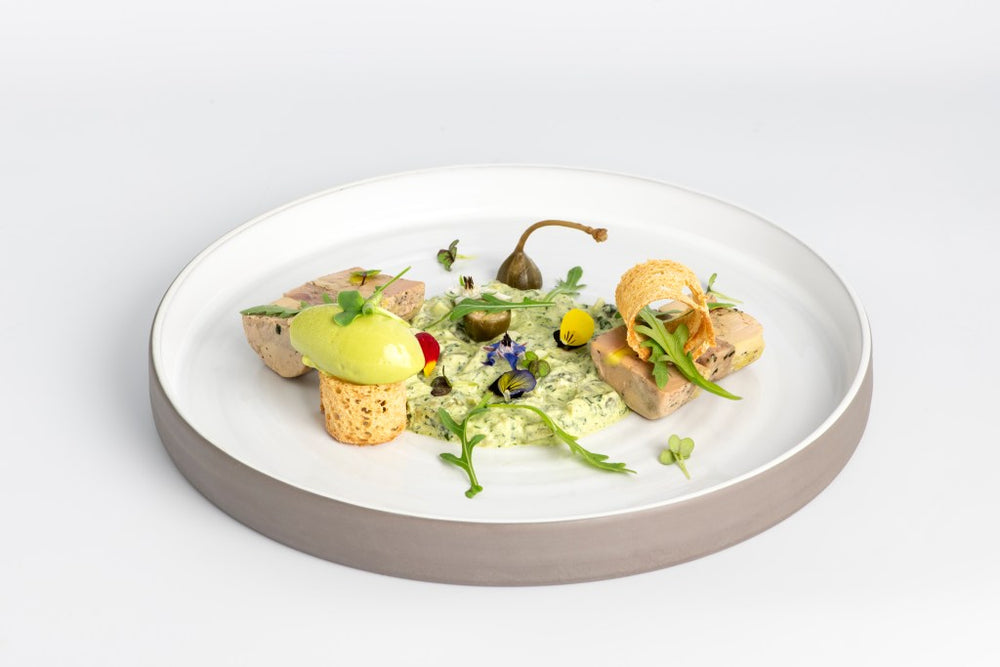 Assiette gastronomique comprenant diverses préparations élégamment disposées avec une mousse verte, des caprons, des fleurs comestibles, et des garnitures colorées.