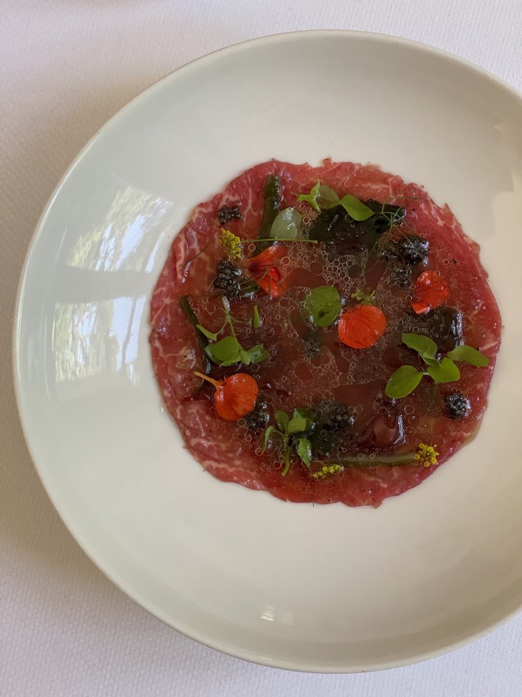 Carpaccio de bœuf garni de petites feuilles et de fleurs comestibles, disposé sur une grande assiette blanche.