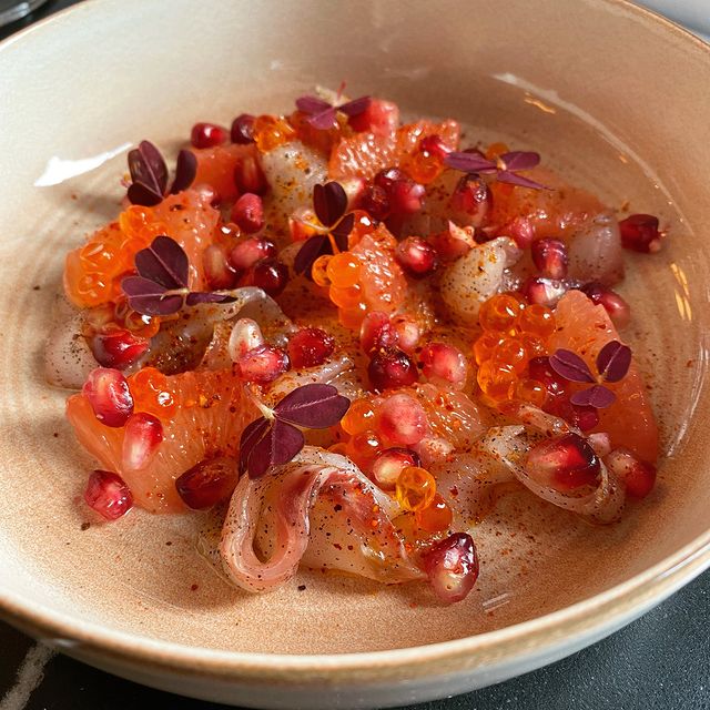 Tartare de poisson agrémenté de grains de grenade, d'œufs de poisson et de feuilles violettes, servi dans un bol en céramique beige.
