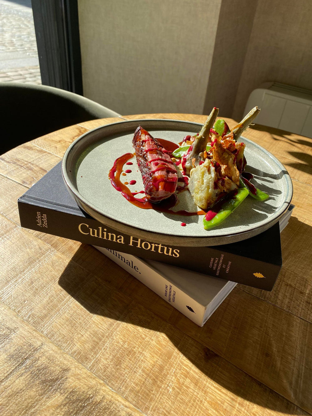 Plat gastronomique dressé sur une assiette, posée sur deux livres empilés intitulés 'Culina Hortus' et 'Animale', avec une sauce rouge éclatante et des légumes artistiquement présentés.