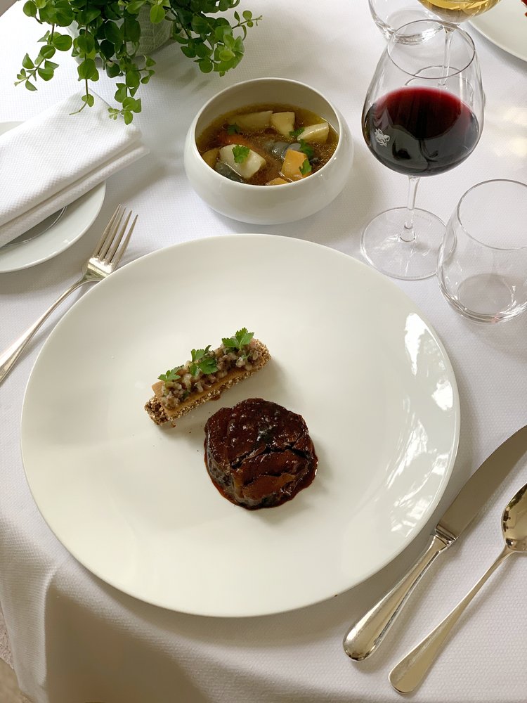 Plat de viande avec une sauce brune, accompagné d'une garniture croquante et d'un bol de soupe en arrière-plan, sur une table blanche élégamment dressée avec un verre de vin rouge.