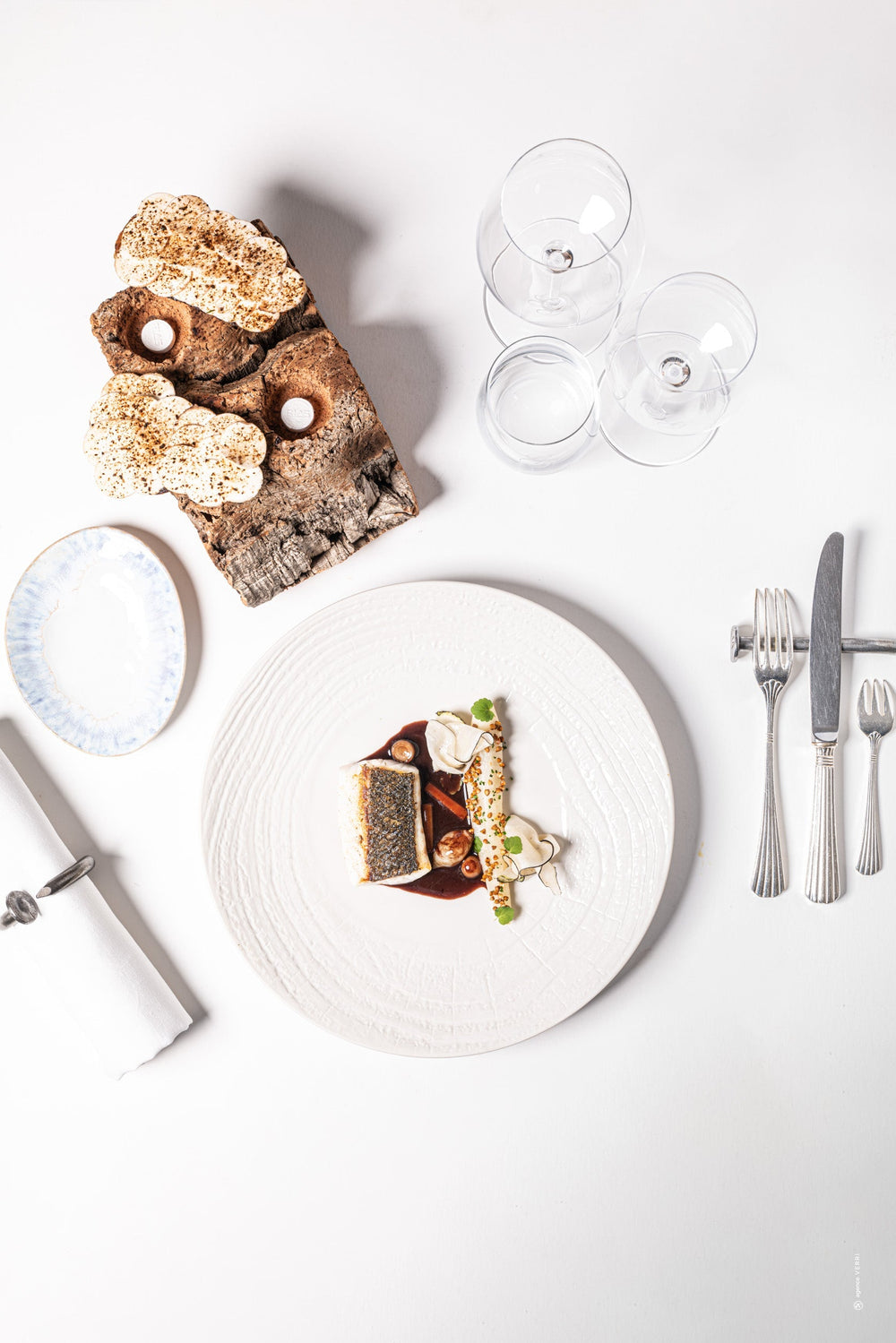 Assiette élégante avec une variété de petits plats gastronomiques, accompagnée de couverts et de verres à vin sur une table blanche.