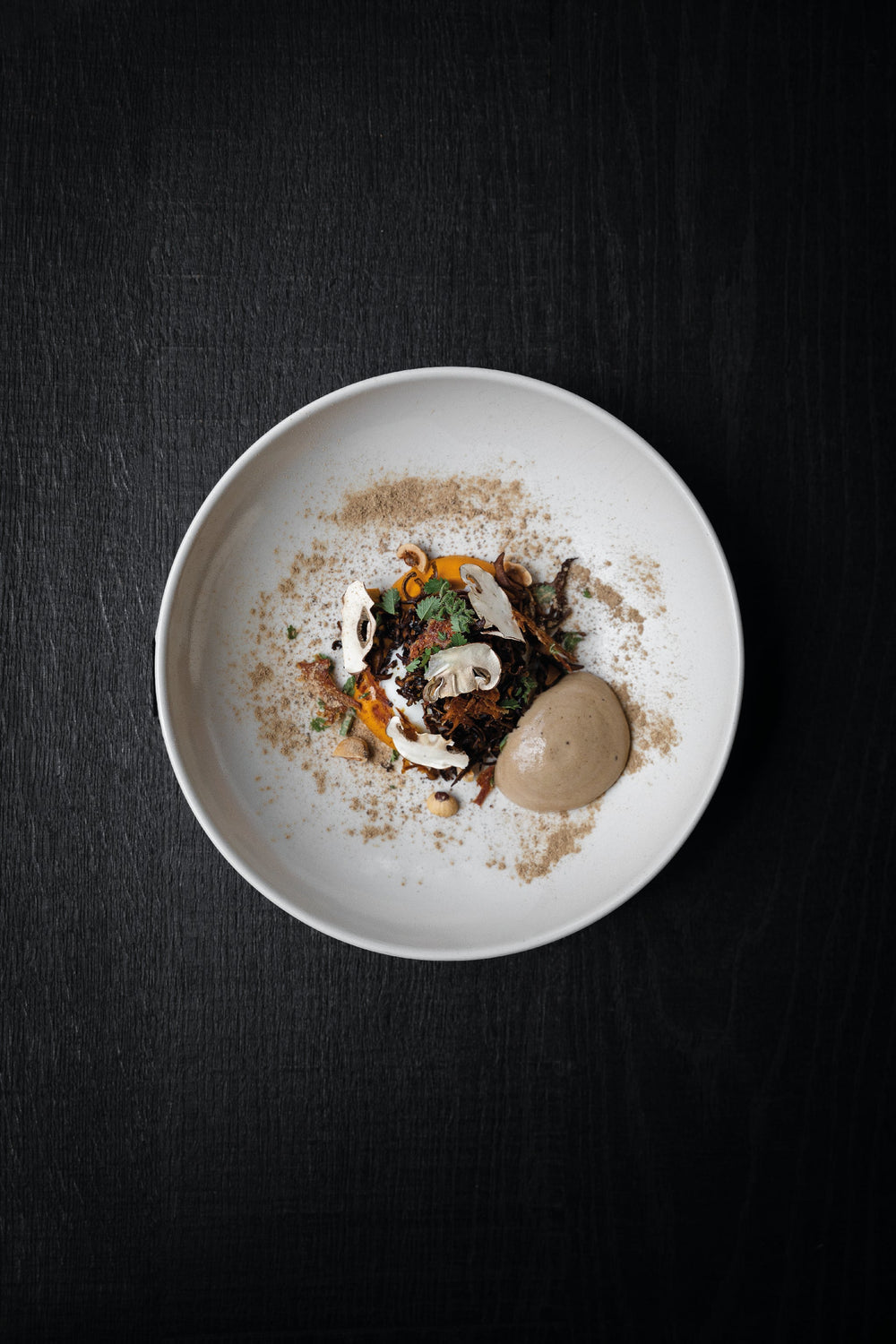 Plat gastronomique élégant présenté dans une assiette blanche, avec des légumes, des champignons et une mousse délicatement disposée, saupoudré d'épices.