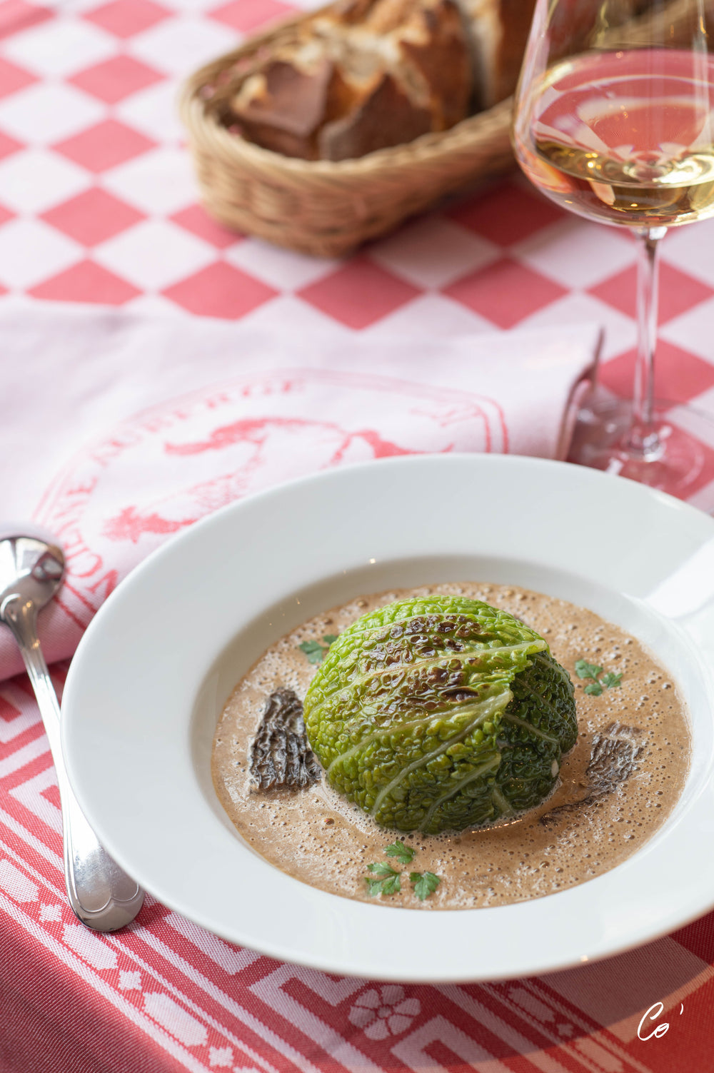 Chou farci servi dans une assiette blanche, accompagné d'une sauce crémeuse et de morilles, posé sur une nappe à carreaux rouges et blancs avec un verre de vin blanc à côté.