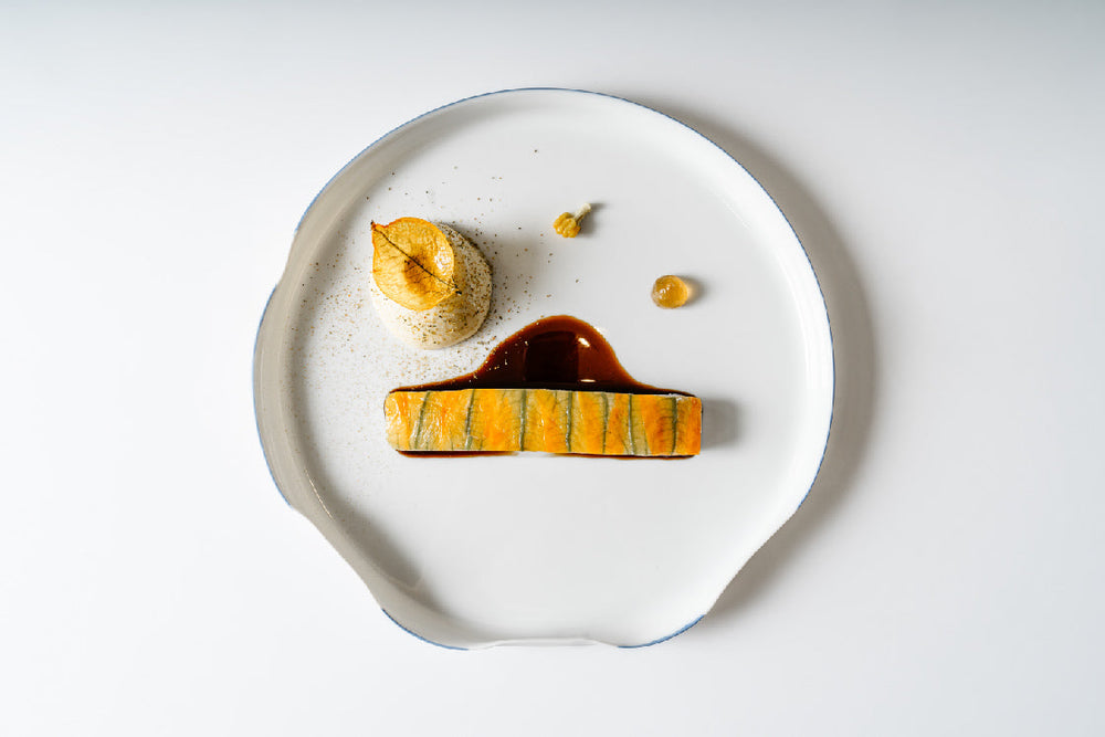 Présentation soignée d'un plat gastronomique avec un filet de poisson mariné, une sauce délicate et des accompagnements élégants, disposé sur une assiette blanche minimaliste.