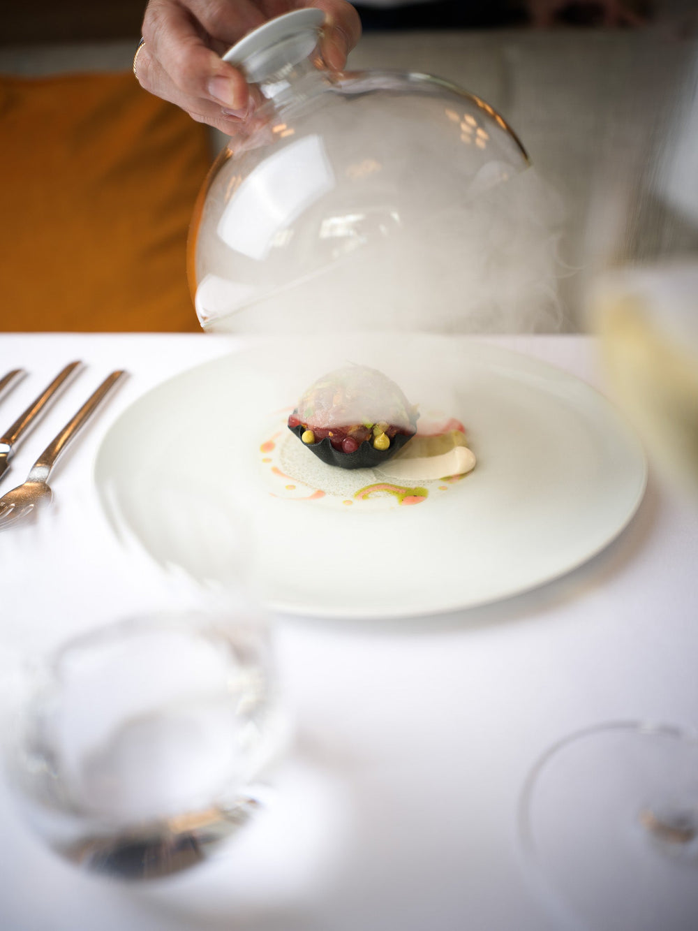 Un plat gastronomique servi sous une cloche en verre, dévoilant une préparation délicate avec des touches de couleur, entouré de vapeur pour un effet spectaculaire.