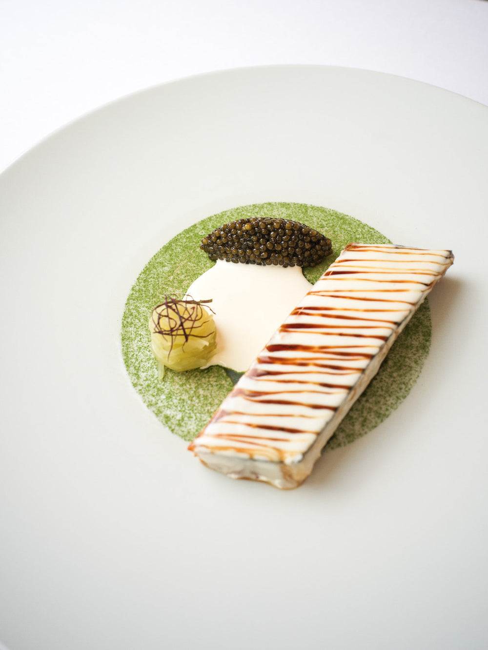 Élégant plat gastronomique avec une barre de poisson nappée de sauce, servie avec du caviar, une crème légère et une garniture délicate sur une assiette décorée de poudre verte.