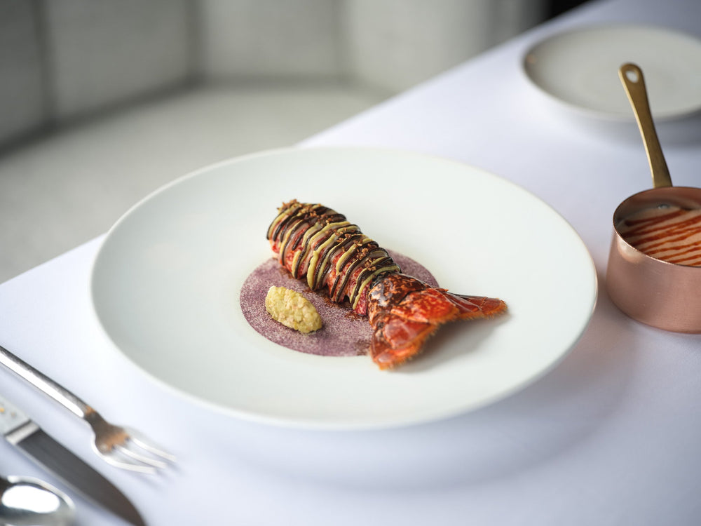 Plat gastronomique composé d'une queue de homard finement dressée avec des légumes en fines lamelles sur une sauce colorée, servi dans une assiette élégante avec une petite casserole en cuivre contenant une sauce à côté.
