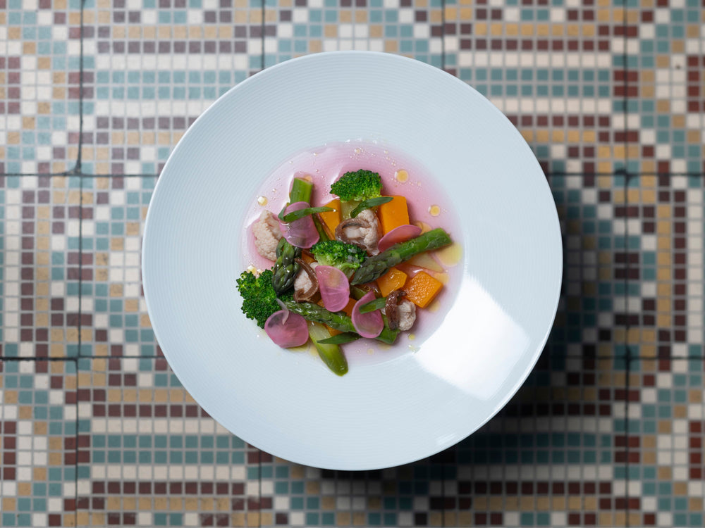 Plat gastronomique coloré présenté dans une assiette blanche, avec des légumes variés tels que des asperges, brocolis, radis et morceaux de potiron, sur un fond de sauce rose. Le tout est posé sur un sol en mosaïque aux motifs géométriques.