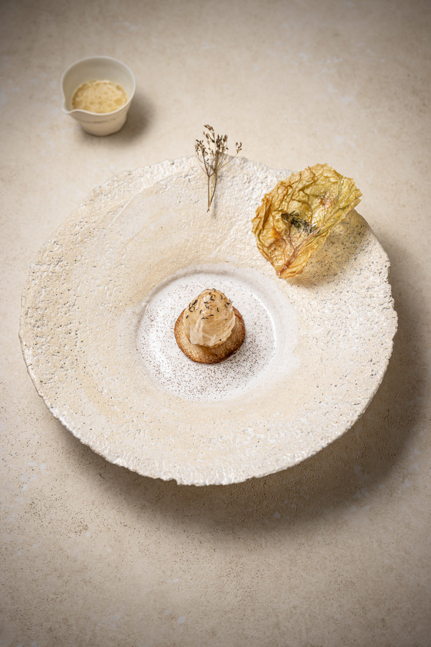 Un plat gastronomique délicatement présenté sur une grande assiette texturée de couleur beige. Le plat principal est une création culinaire artistique garnie d'une feuille croquante et d'une petite plante décorative, avec une sauce à côté dans une petite tasse blanche.