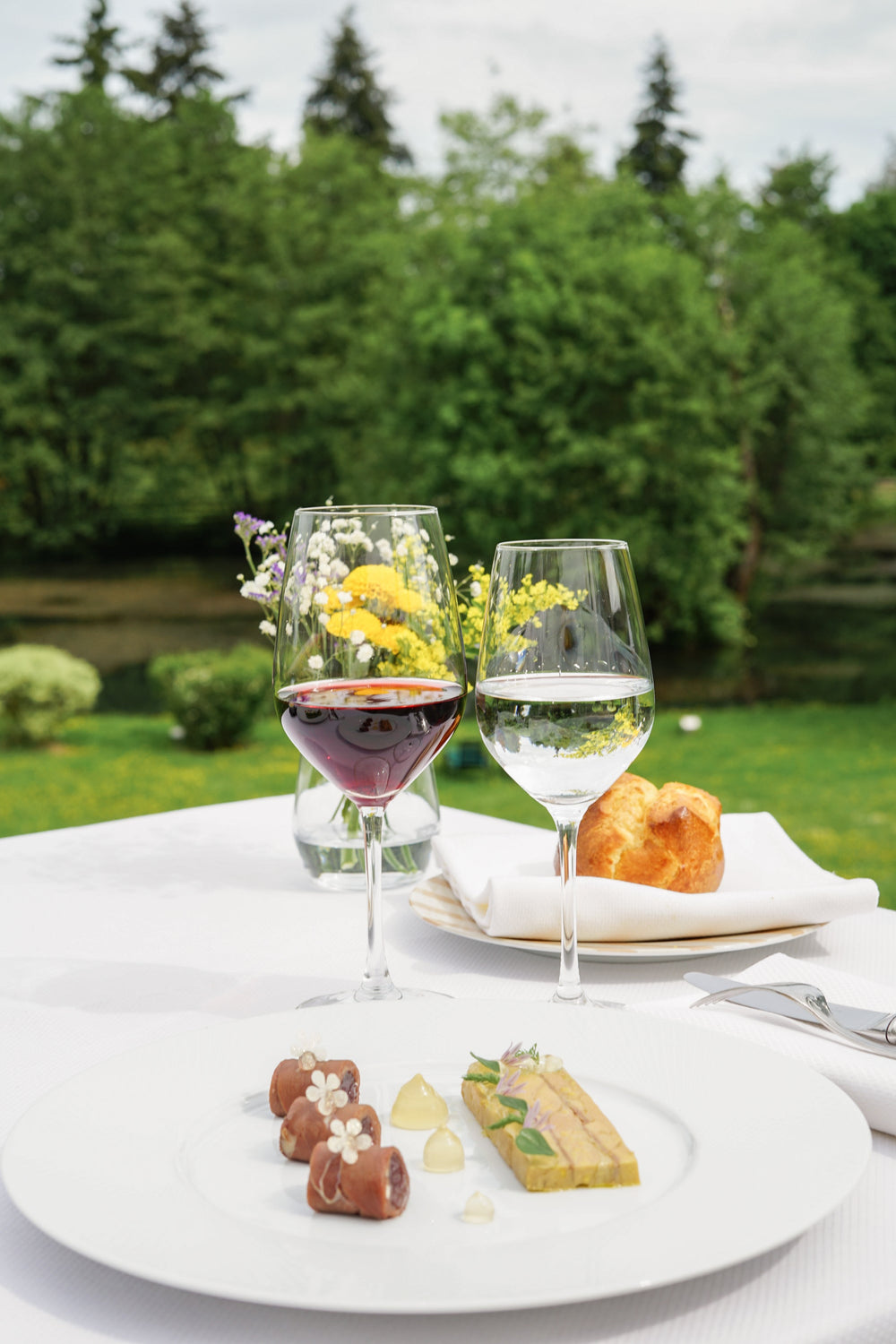 Un déjeuner gastronomique en plein air avec une assiette élégante de mets raffinés, deux verres de vin, et un bouquet de fleurs sauvages en arrière-plan, sur fond de verdure luxuriante.