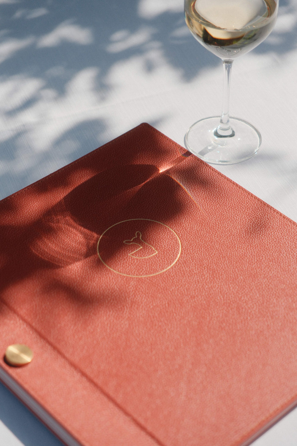 Menu de restaurant en cuir rouge avec un logo doré en relief, posé sur une table élégante avec un verre de vin blanc, sous un jeu d'ombres naturelles projetées par la lumière.
