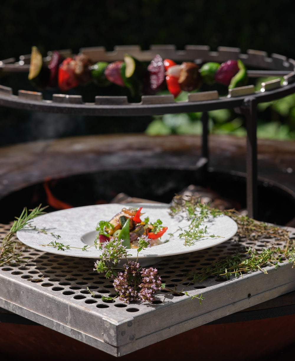 Assiette de légumes grillés présentée devant un barbecue avec des brochettes de légumes en train de cuire, décorée de plantes aromatiques.