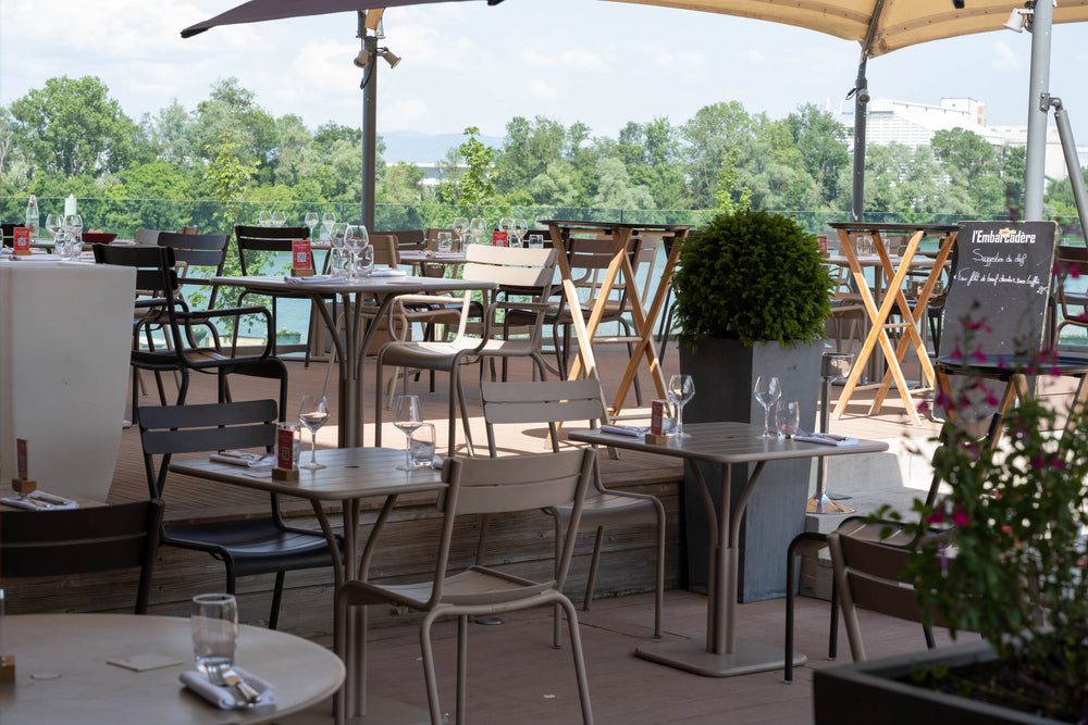 Terrasse de restaurant élégante avec vue sur une rivière bordée de verdure, tables dressées avec verrerie et ardoise de menu à l'entrée.