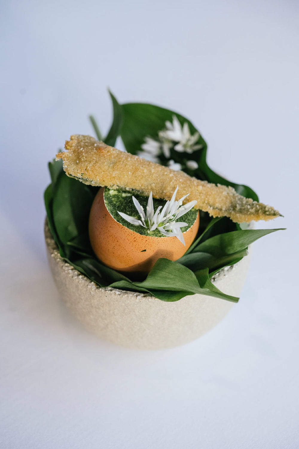Un œuf servi dans sa coquille, garni de mousse verte et décoré de petites fleurs blanches, accompagné d'un bâtonnet croustillant, le tout présenté sur un lit de feuilles vertes dans un bol élégant.