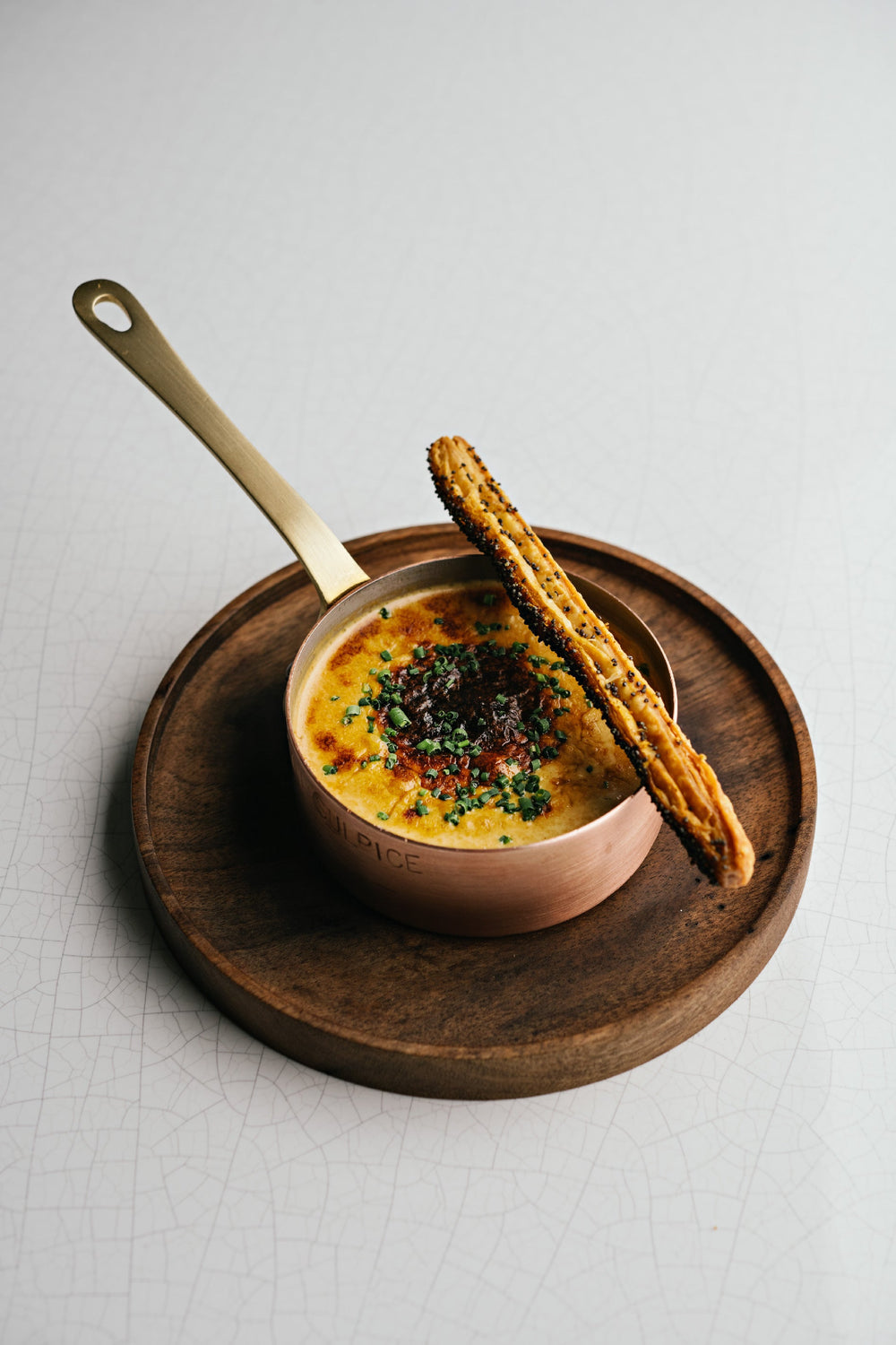 Un plat gastronomique servi dans une casserole en cuivre, garni de fines herbes et accompagné d'un bâtonnet feuilleté doré, présenté sur une assiette en bois.