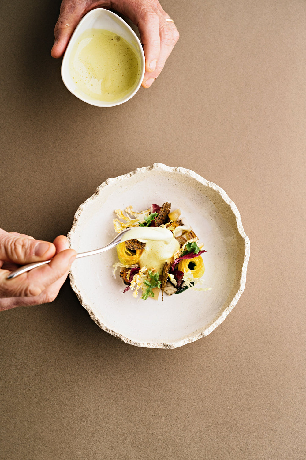 Un plat raffiné dans une assiette en céramique texturée, garni de légumes délicatement disposés, tandis qu'une main ajoute une sauce crémeuse depuis une cuillère, apportant la touche finale à cette présentation gastronomique.