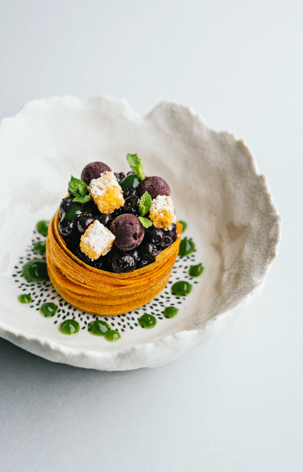 Dessert raffiné présenté dans une assiette en céramique, composé d'une base croustillante garnie de petites baies, de cubes de garniture dorée et saupoudrée de sucre, décorée de feuilles de menthe et entourée de points de coulis vert.