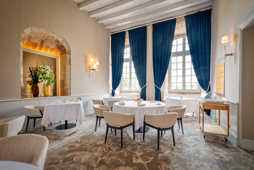 Salle de restaurant lumineuse et élégante avec des tables rondes et carrées dressées de nappes blanches, des chaises rembourrées confortables, des grandes fenêtres encadrées de rideaux bleus et une décoration florale raffinée.