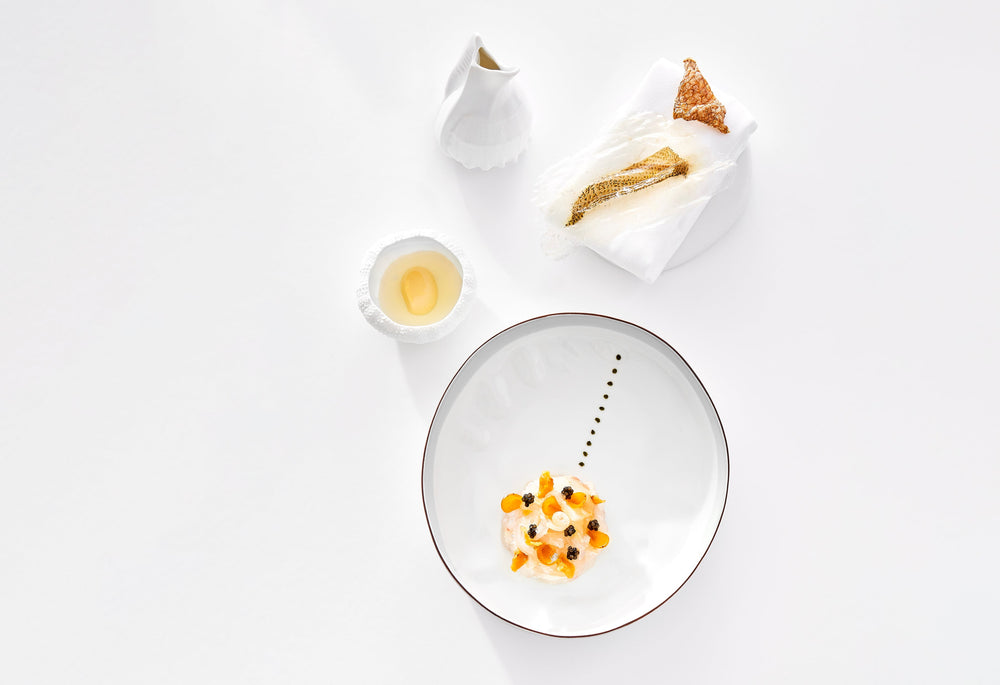 Plat gastronomique épuré avec une présentation minimaliste, composé de poisson délicatement dressé, accompagné de petits condiments et d'une sauce légère, sur une assiette blanche avec un motif subtil.