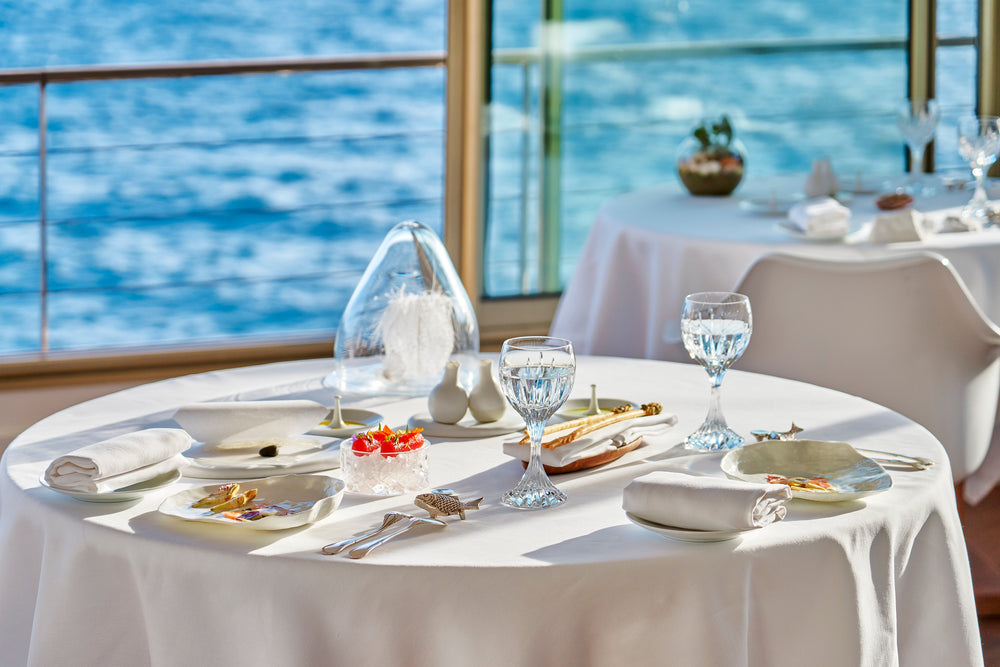 Table élégamment dressée avec vue sur la mer, dans un restaurant gastronomique, ornée de verres en cristal et de plats raffinés, créant une atmosphère de luxe et de sérénité.