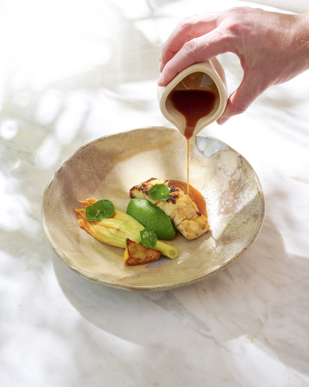 Main versant une sauce sur un plat composé de poisson, de légumes et de purée verte.