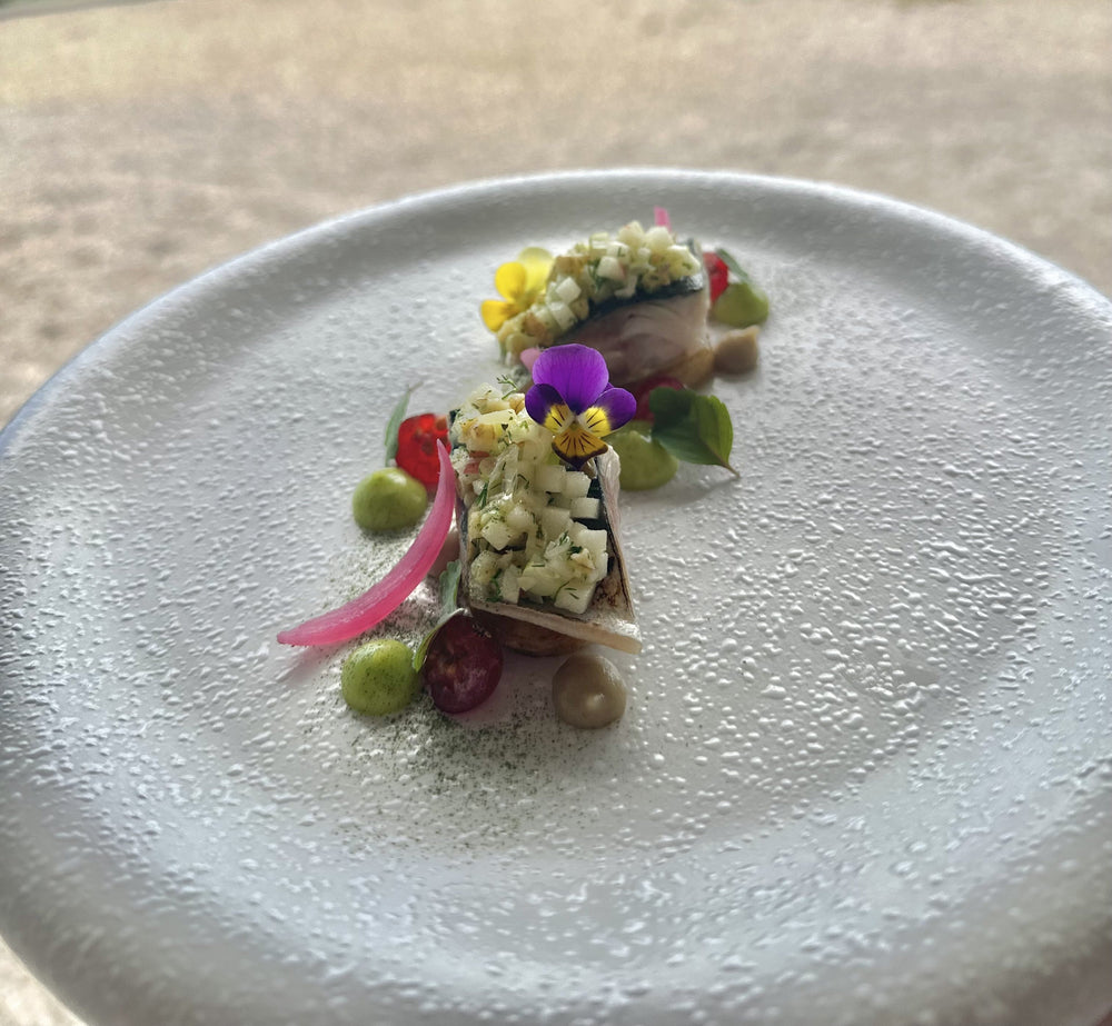 Une assiette gastronomique délicatement présentée, comprenant des morceaux de poisson garnis de dés de légumes, des pickles d'oignon rouge et des petites touches de purée colorée, le tout décoré de fleurs comestibles et de feuilles fraîches, sur une assiette texturée blanche.