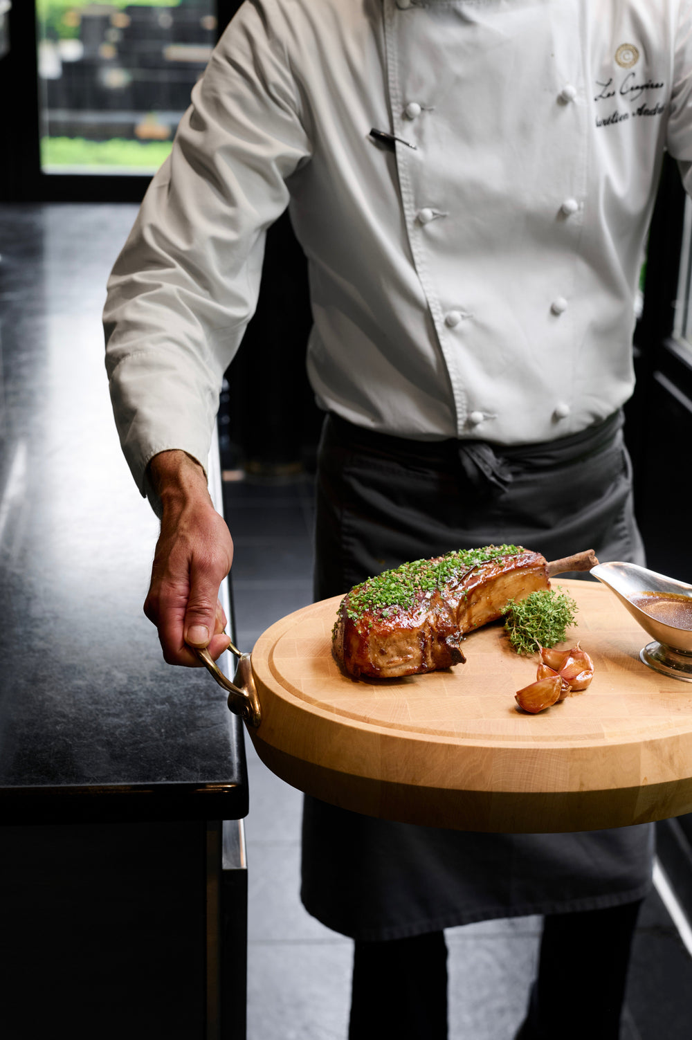 Chef tenant une planche de bois avec une côte de porc garnie d'herbes et des gousses d'ail rôties