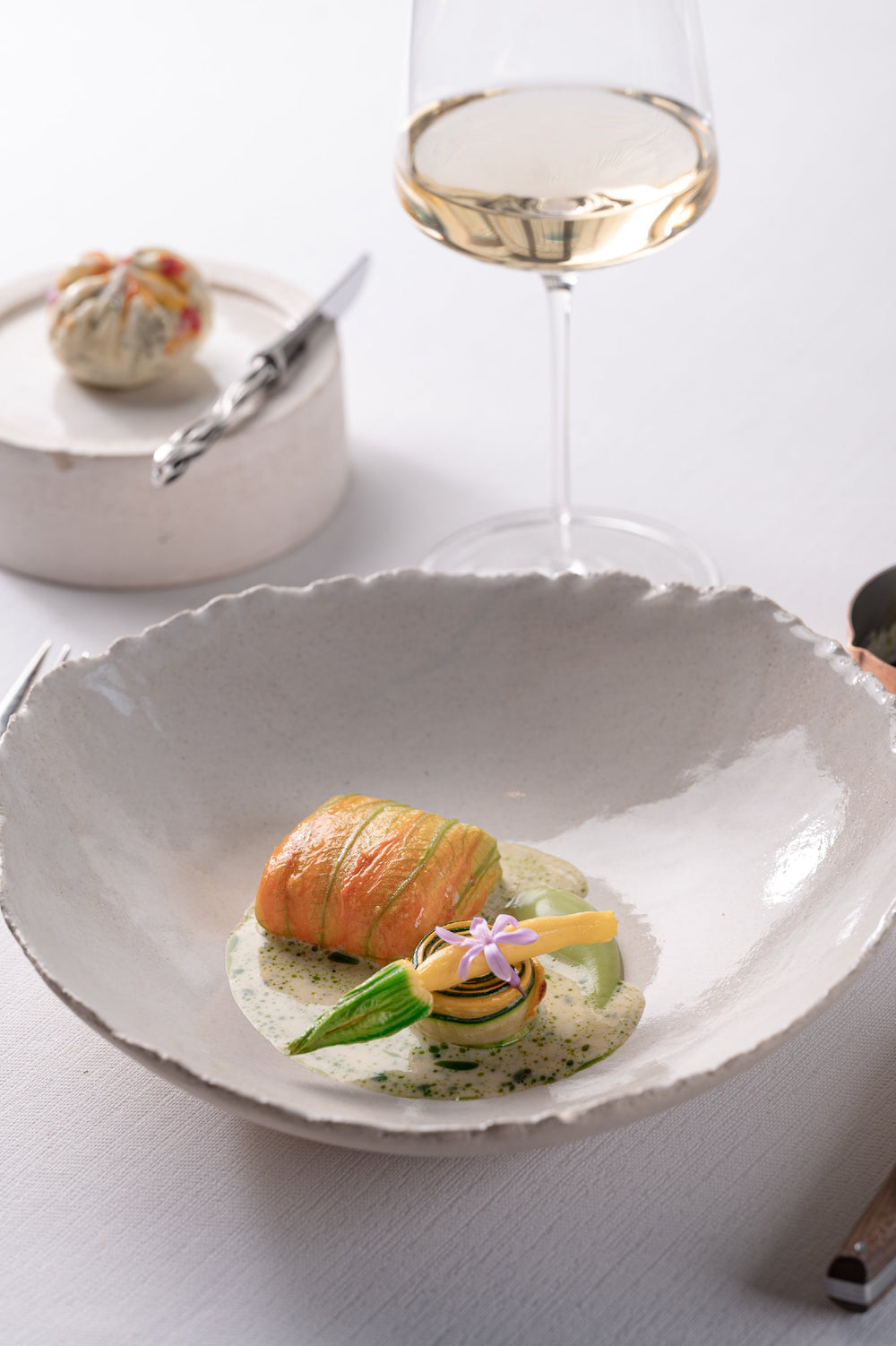 Plat gastronomique composé d'un roulé de légumes délicatement dressé dans une assiette en céramique, accompagné d'une sauce onctueuse et décoré d'une fleur comestible, avec un verre de vin blanc en arrière-plan.