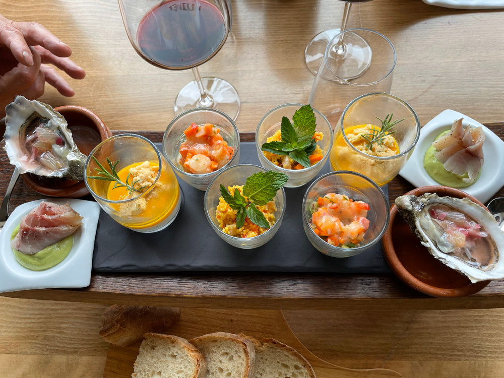 Plateau d'entrées gastronomiques composé de verrines variées, huîtres, tranches de poisson fumé et autres délicatesses, accompagné de pain et d'un verre de vin rouge.
