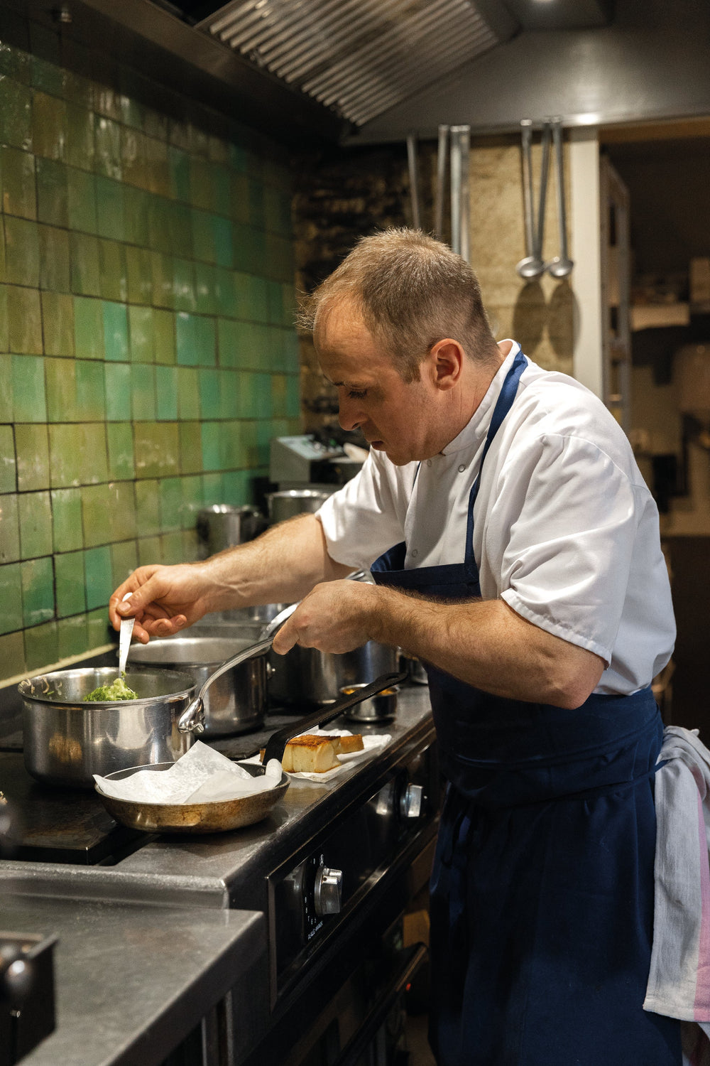Un chef cuisinier concentré en train de préparer un plat dans une cuisine professionnelle avec des carreaux verts en arrière-plan.