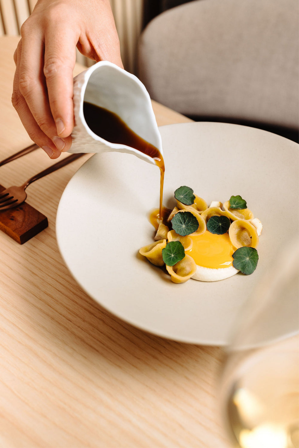Plat gastronomique de pâtes artisanales garnies de feuilles de capucine, avec une sauce brune versée délicatement depuis une petite carafe blanche, présenté sur une table en bois clair.