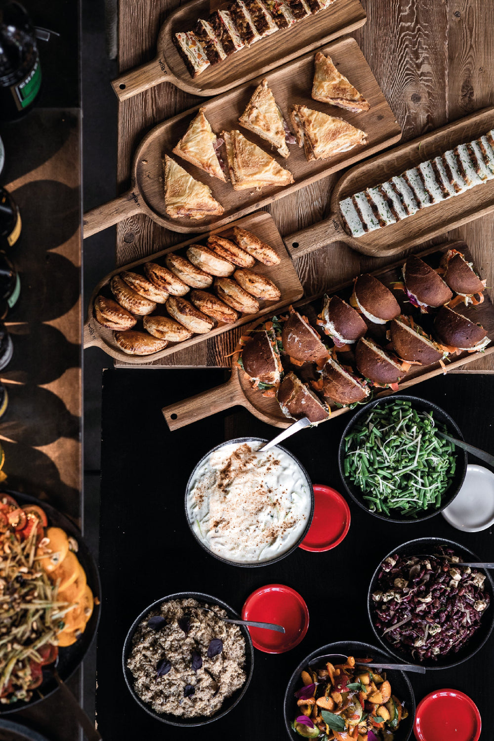 Vue aérienne d'un buffet varié comprenant des sandwichs, des feuilletés, des pâtisseries salées et des bols de salades et de sauces, disposés sur des planches en bois.