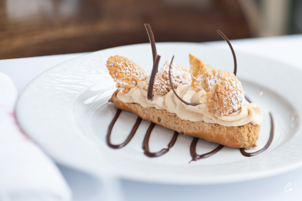 Éclair délicatement présenté, garni de crème légère et décoré de motifs en chocolat et de sucre glace, sur une assiette blanche.