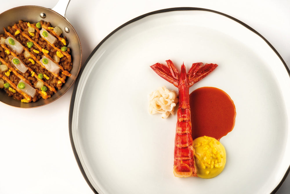 Assiette gastronomique composée d'une crevette rouge dressée avec une sauce brune, une mousse jaune, et des nouilles, accompagnée d'une poêle de paëlla garnie de légumes et de poisson.