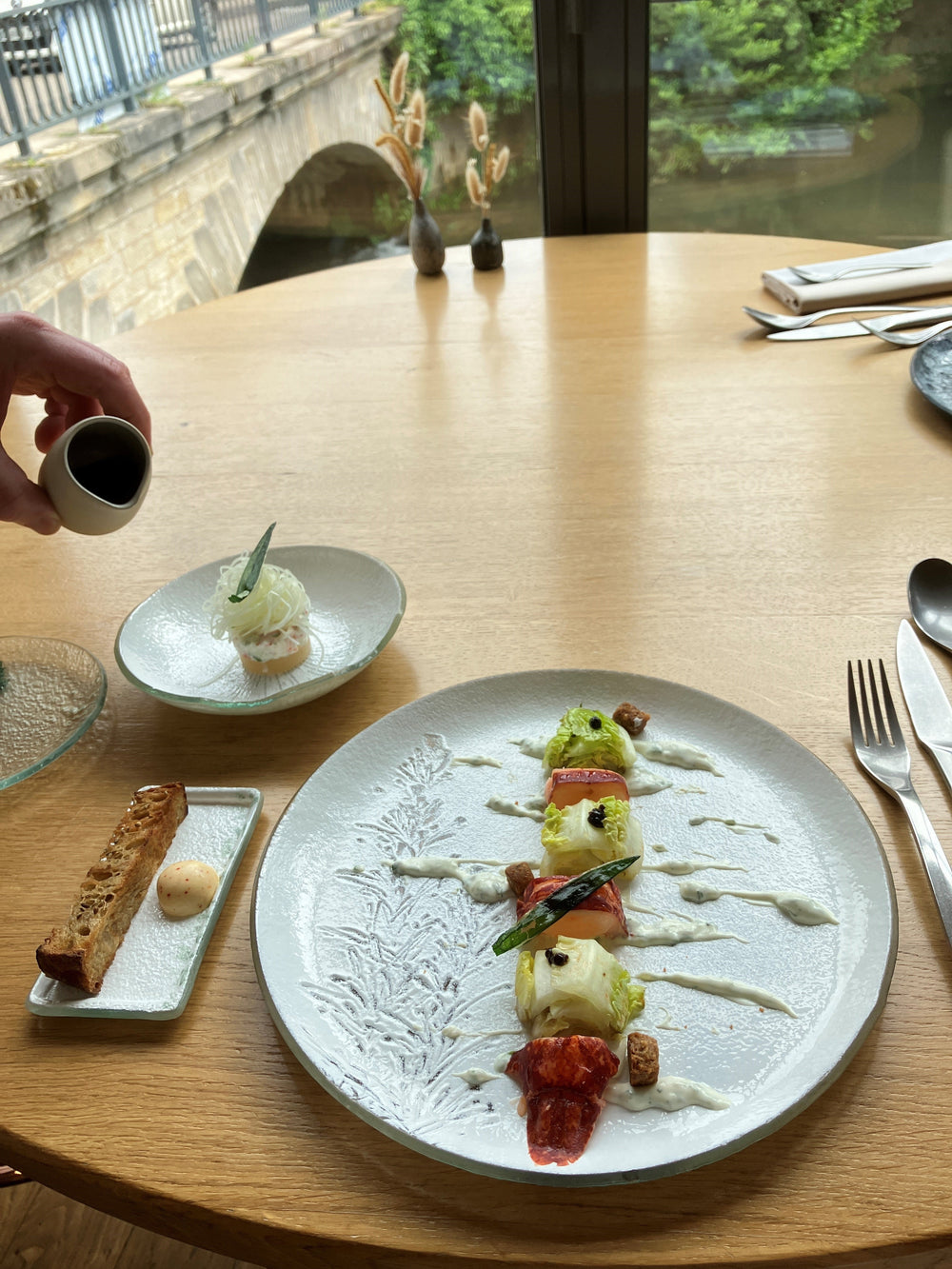 Assiette gastronomique élégante avec des morceaux de homard et de laitue, joliment disposés avec une sauce légère, accompagnée de pain grillé et d'une vue sur un pont et la nature en arrière-plan.