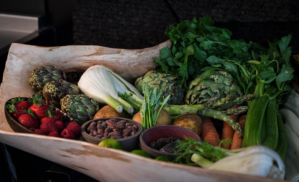 Un panier en bois rempli de légumes frais, dont des artichauts, du fenouil, des asperges, des carottes, des pommes de terre et des céleris, ainsi que des fraises et des framboises.