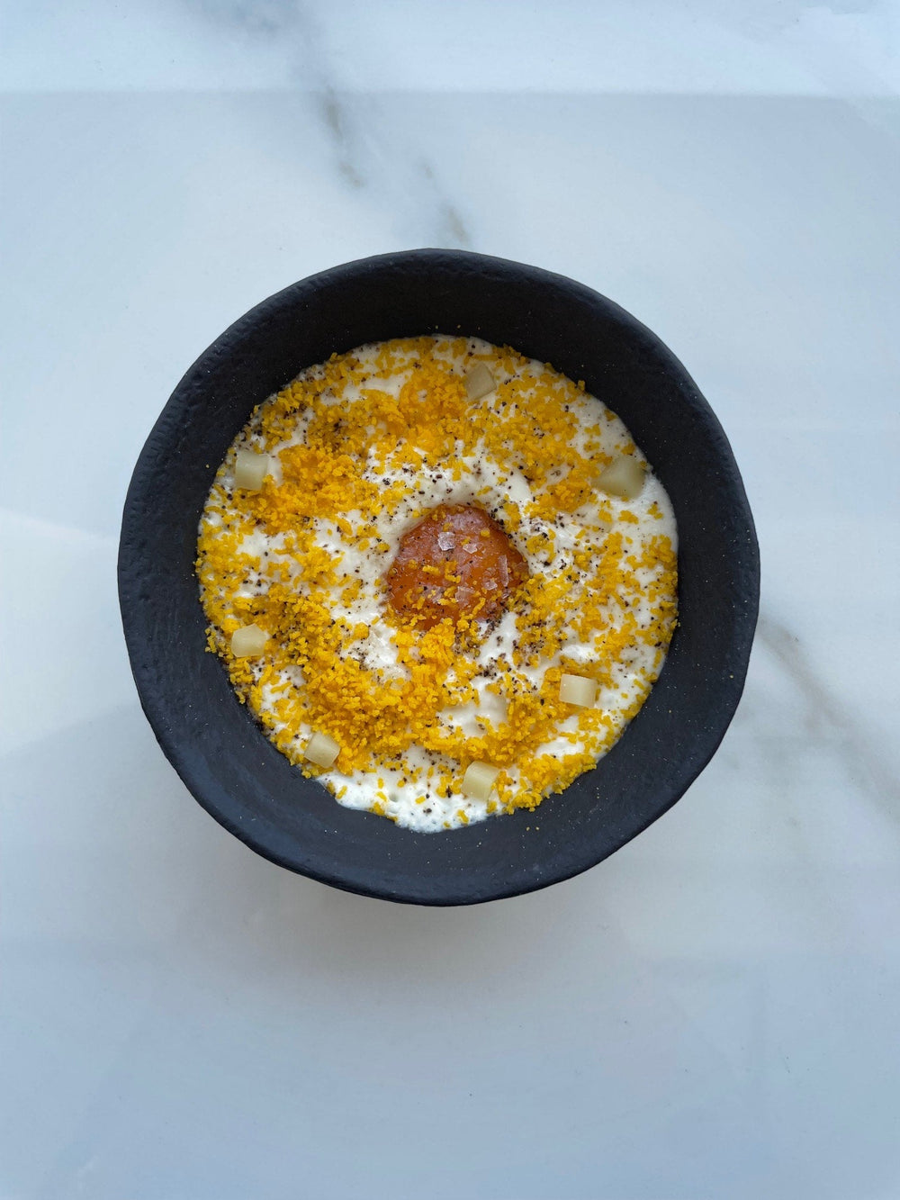 Un plat gastronomique composé d'un œuf poché au centre, entouré de morceaux de légumes et saupoudré de miettes dorées dans un bol noir sur une table en marbre blanc.